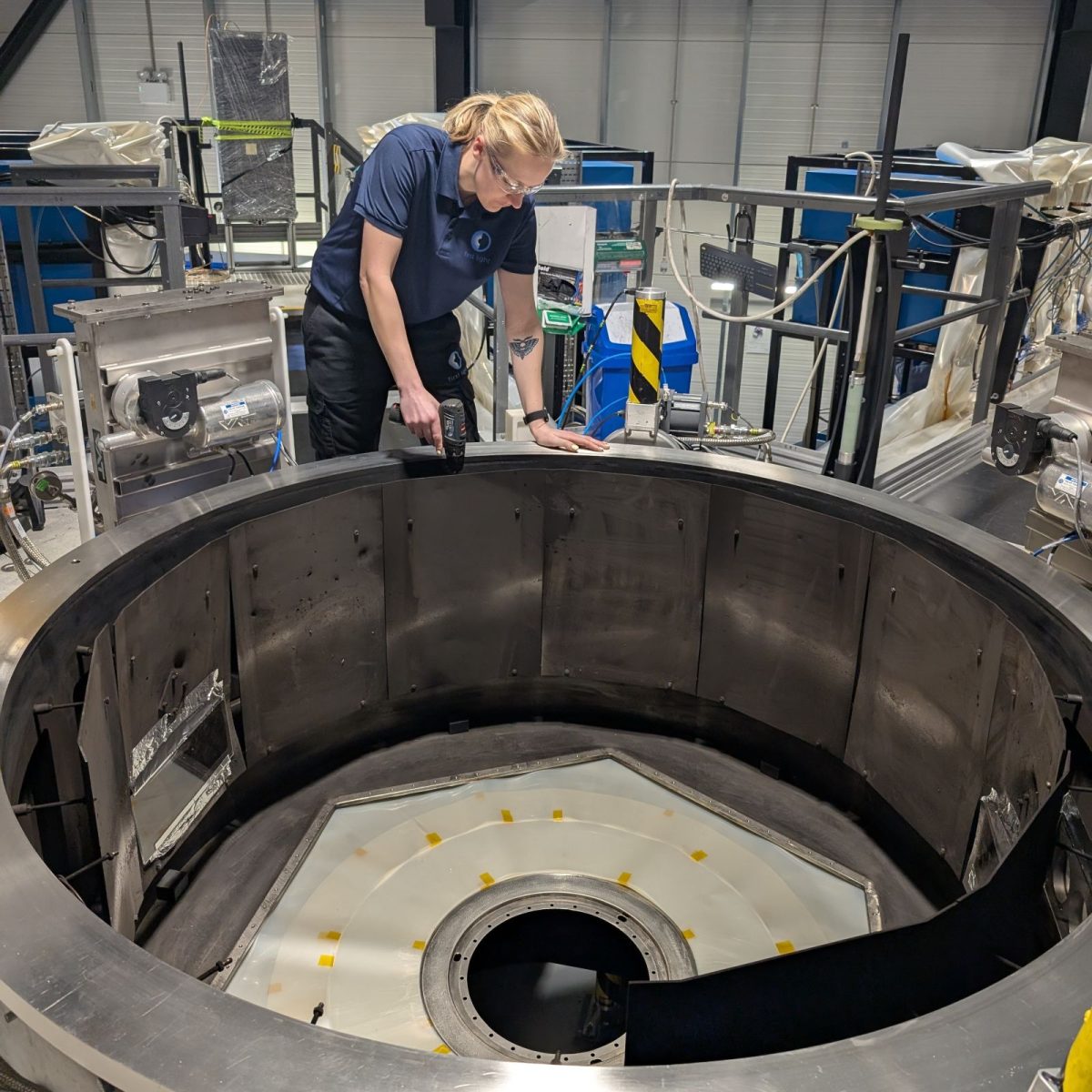 Research and Development technician Kate, in the experimentation area surrounded by equipment, using a drill on a piece of large circular panelling.