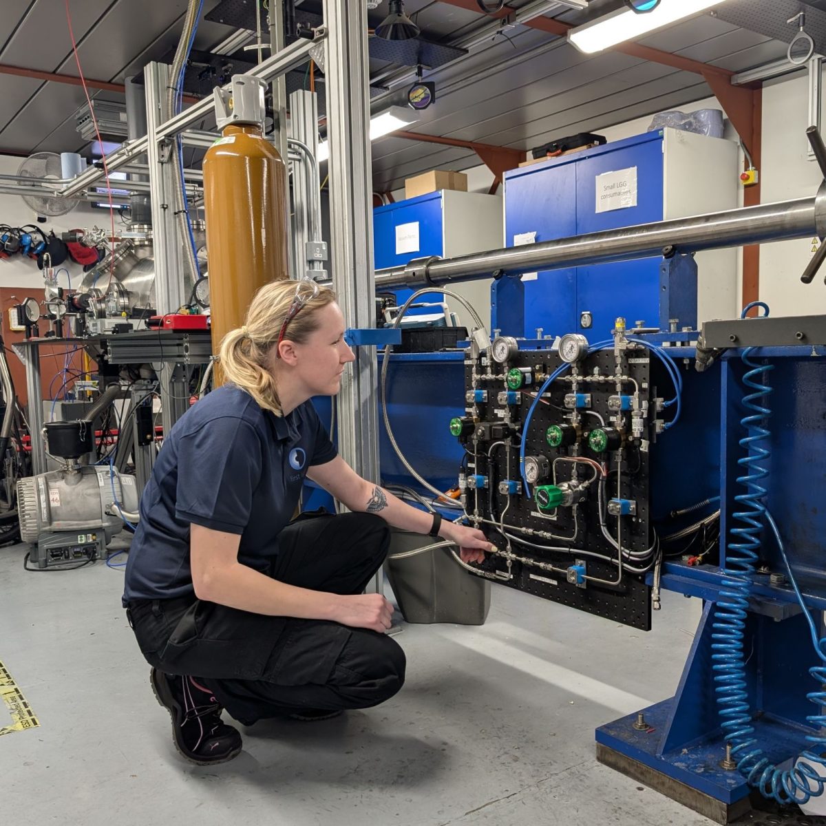 Research and Development technician Kate, in the experimentation area surrounded by equipment, with her safety goggles on her head, crouching next to a panel of gas tubes and dials.