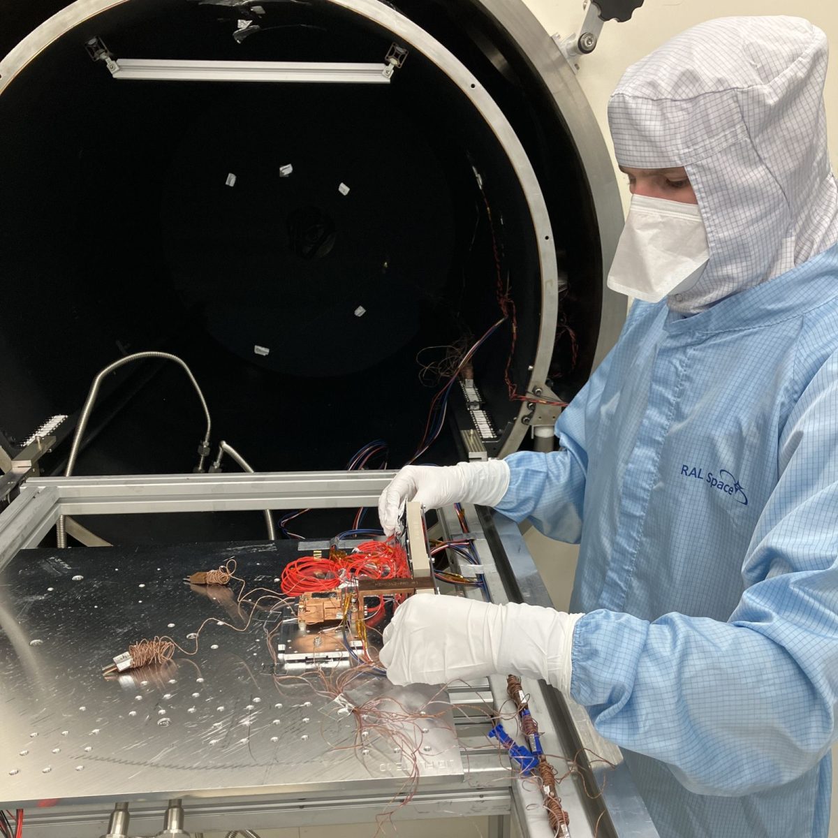 Space Engineering Technician Reuben wearing a full clean suit, putting together electronic components on a worktable.