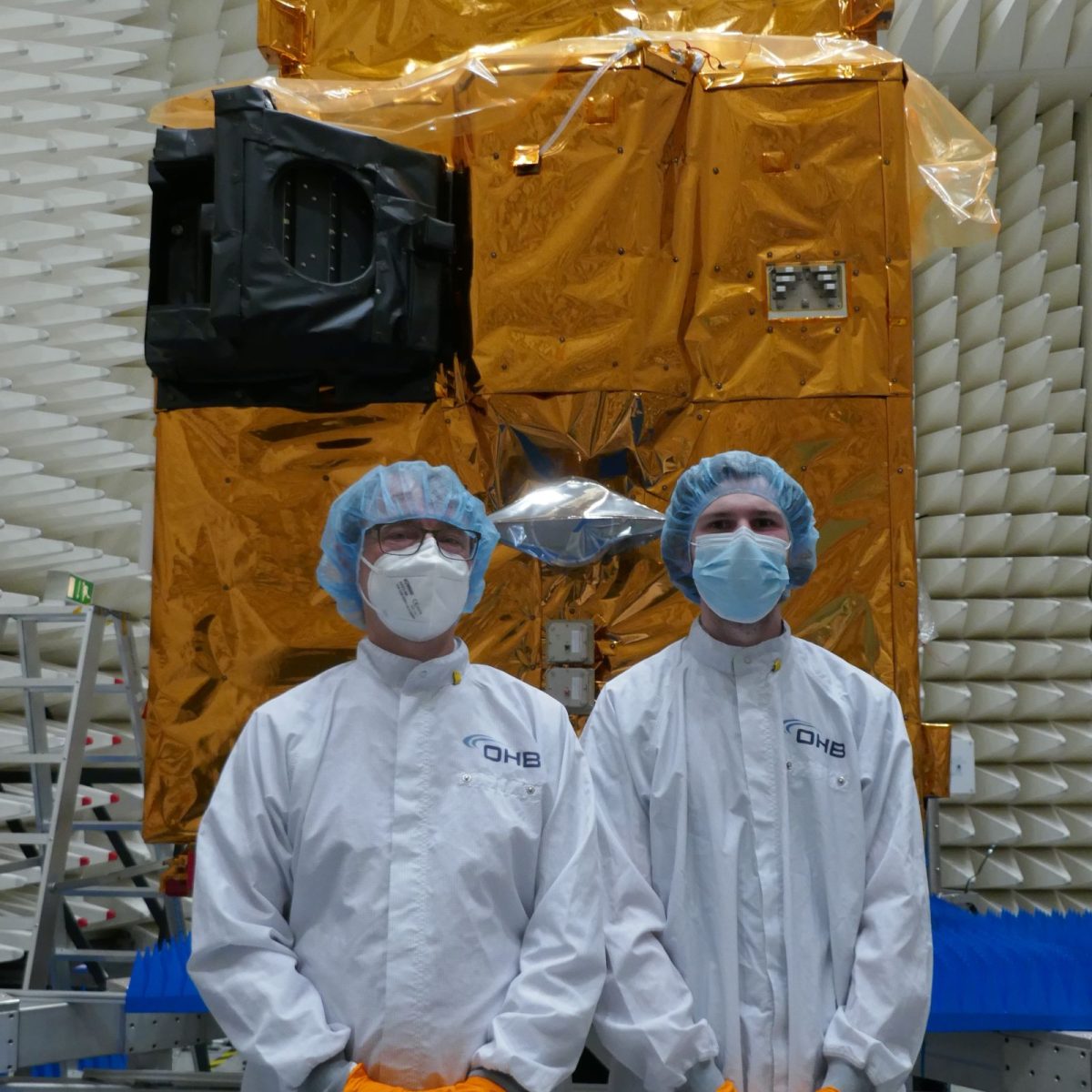 Space Engineering Technician Reuben wearing a full clean suit, standing with a colleague in front of a finished satellite.
