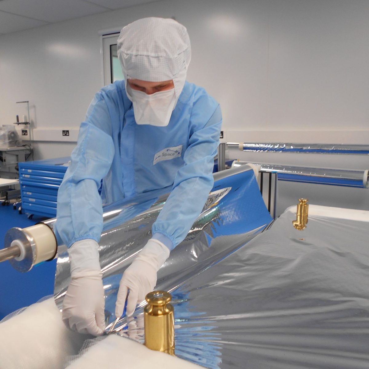 Space Engineering Technician Reuben wearing a full clean suit, measuring out a large roll of aluminium foil.