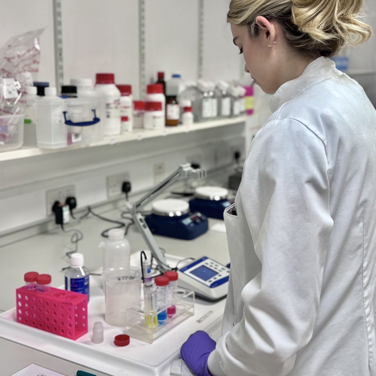 University technician Taylor in the lab, wearing a lab coat and gloves, looking at some test tubes in a rack, and other equipment on the lab desk.