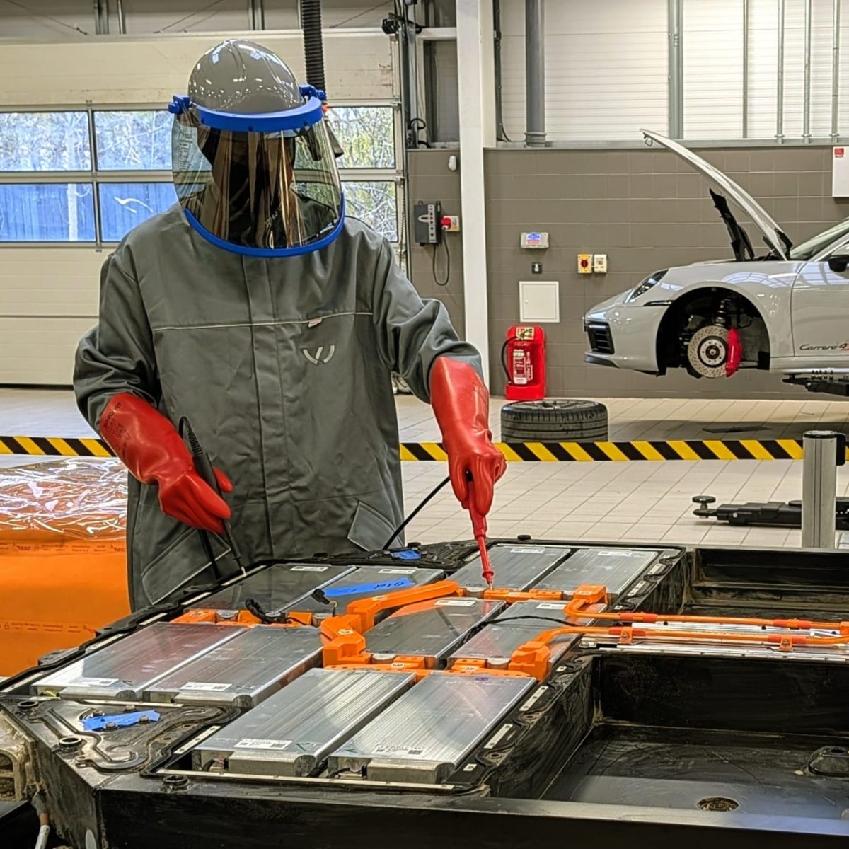 Electric Vehicle Technician Callum, in the garage, working with high voltage equipment on a tabletop, wearing a full arc flash suit, including face visor and insulated rubber gloves.