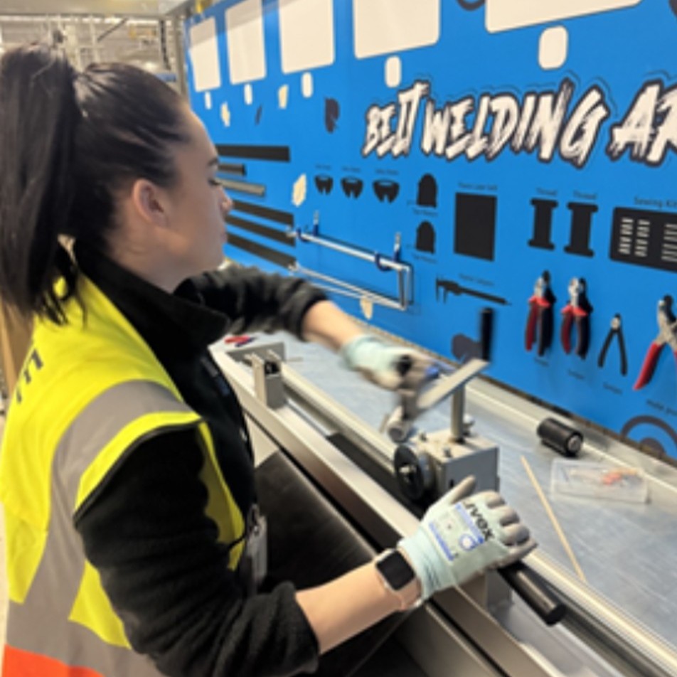 Mechatronics Technician Chloe, wearing a hi-vis vest and gloves using a bench-mounted tool in the workshop.