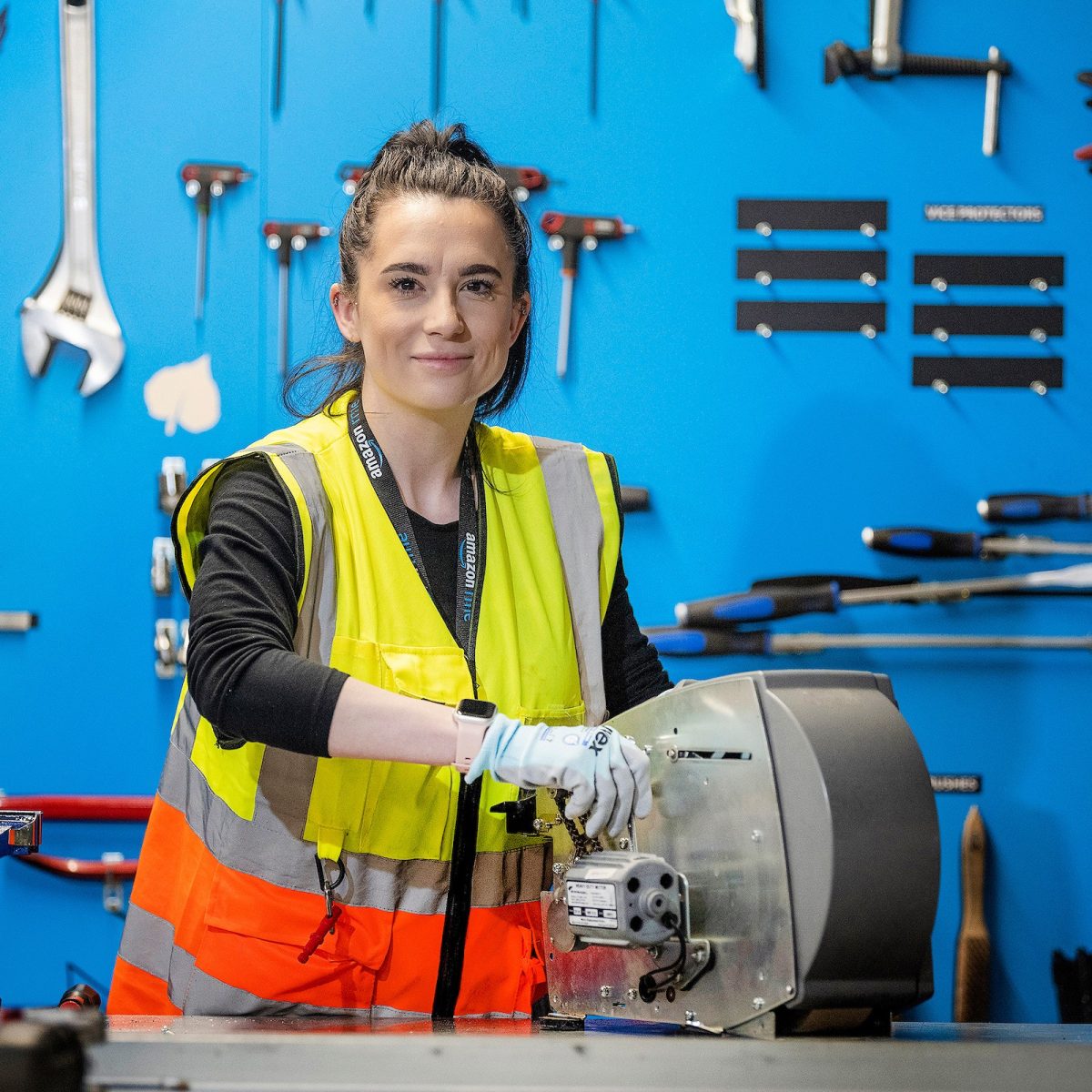 Mechatronics Technician Chloe, in the workshop, wearing a hi-vis vest and gloves, posing by a bench-mounted tool.