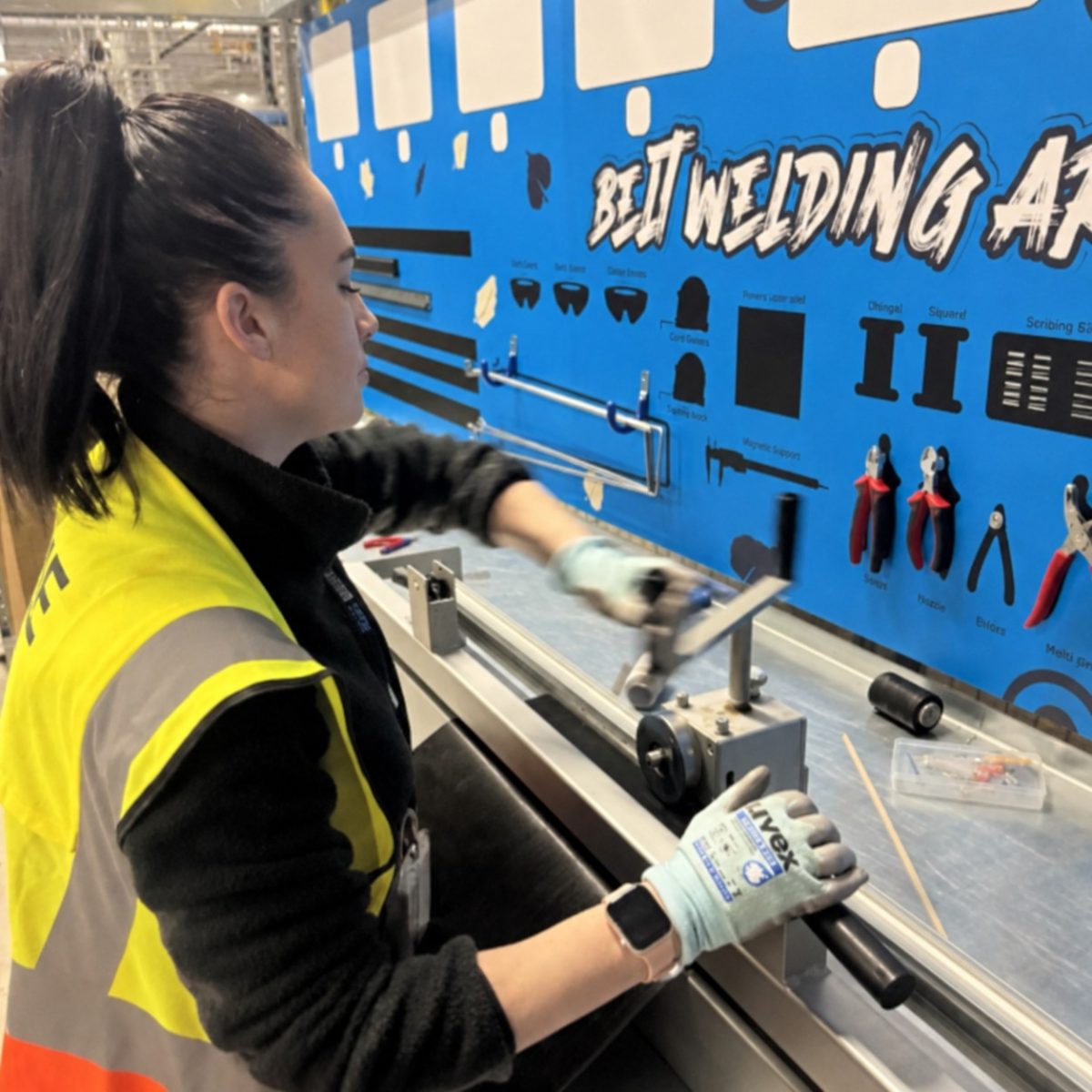 Mechatronics Technician Chloe, wearing a hi-vis vest and gloves using a bench-mounted tool in the workshop.