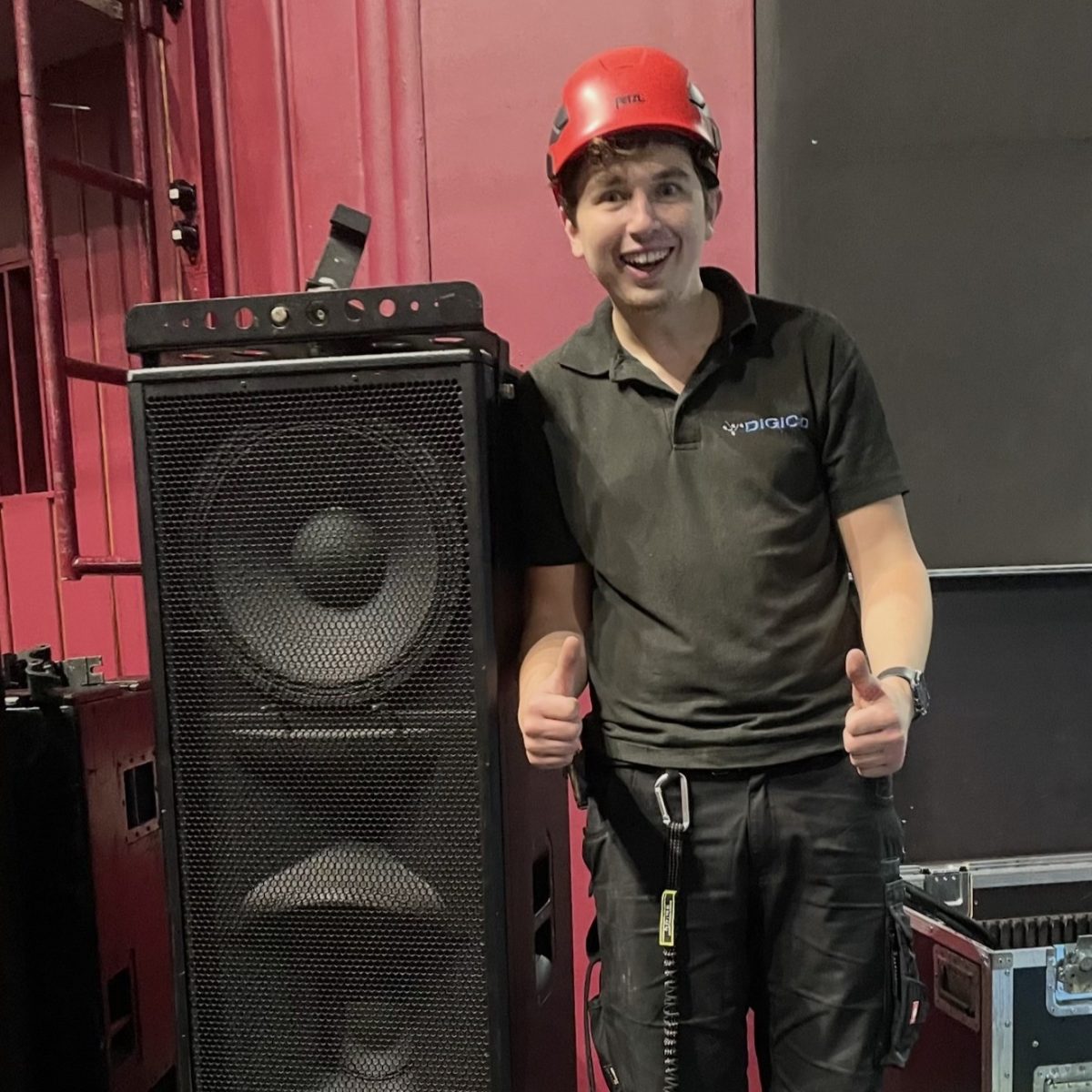 Creative Venue Technician Declan, wearing a protective helmet and harness, smiling and giving the thumbs up, stood next to a large speaker that comes up to this shoulder.