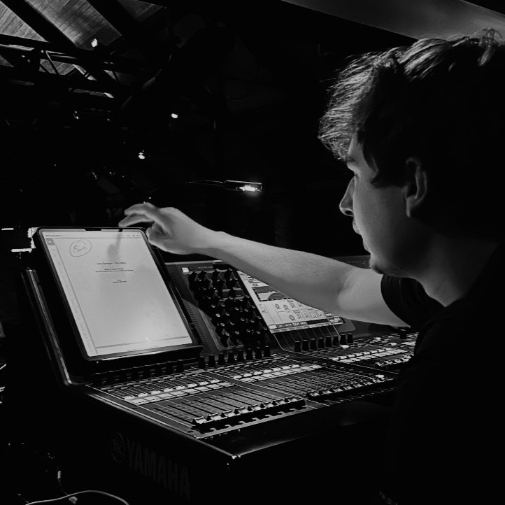 Creative Venue Technician Declan, sat at a lighting mixing desk at the back of a theatre, with a copy of the script, following along and cueing the lights.