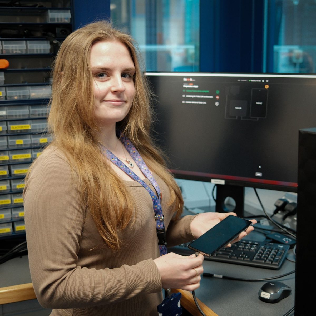 Digital Forensics Technician Elisha, holding a mobile phone, standing in front of a workstation with a computer monitor, keyboard and mouse.