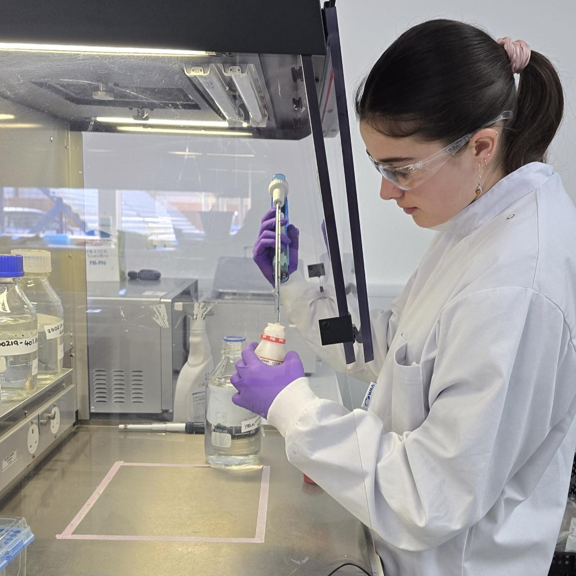 Research Laboratory Technician Eve, in the lab, wearing a lab coat, safety glasses, and latex gloves, working with chemicals in a fume cupboard.