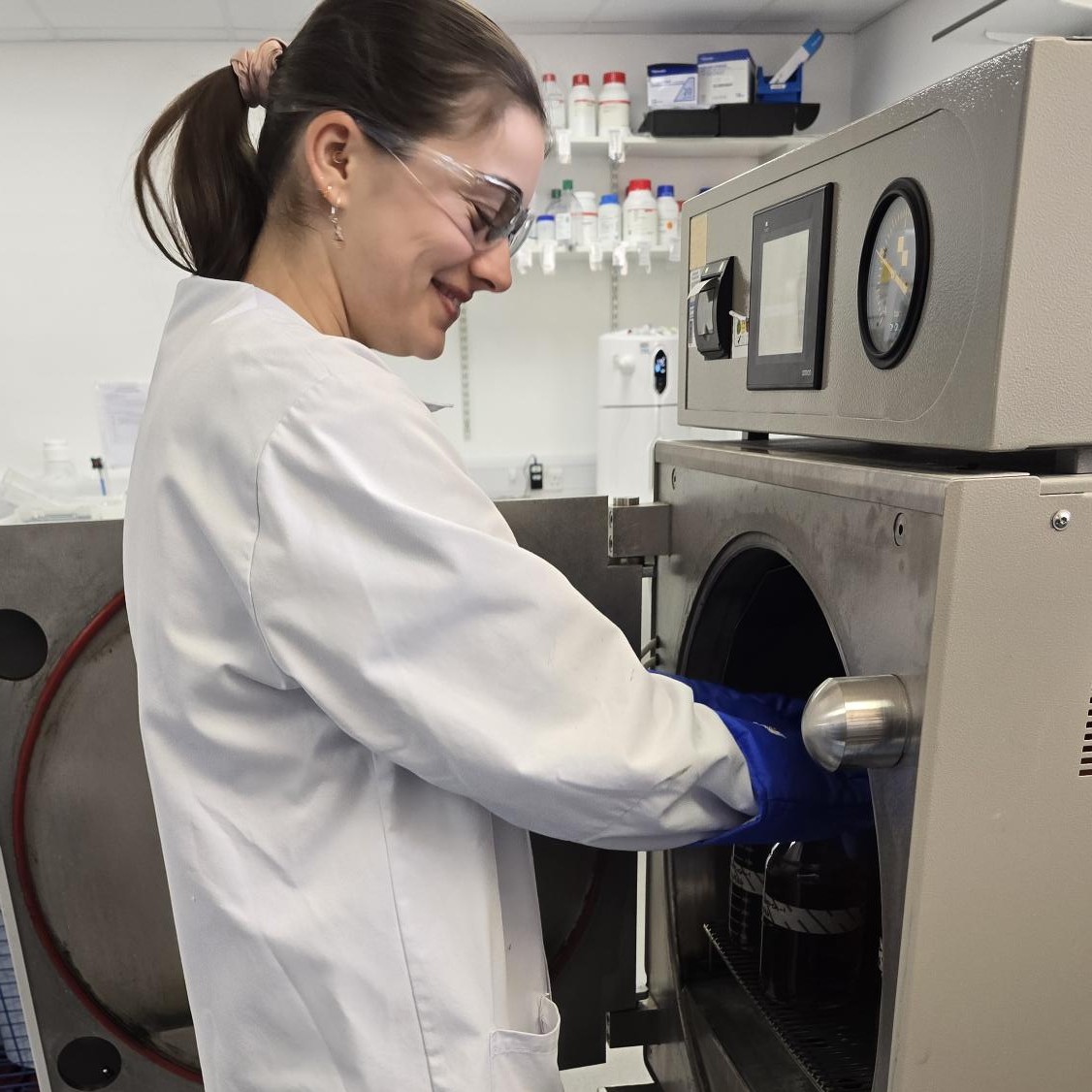 Research Laboratory Technician Eve, in the lab, wearing a lab coat, safety glasses, and thick insulating gloves, putting lab equipment into an autoclave.