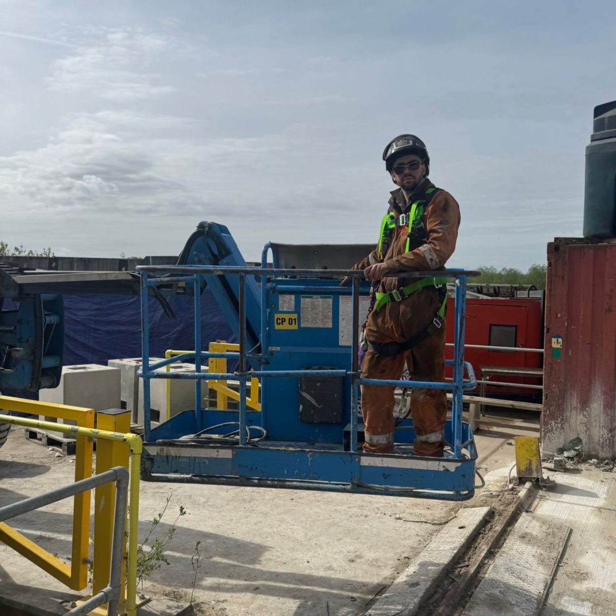Land Based Engineering Technician Finn, standing on a lifting platform in the yard, wearing overalls, protective gloves, a helmet and sunglasses.
