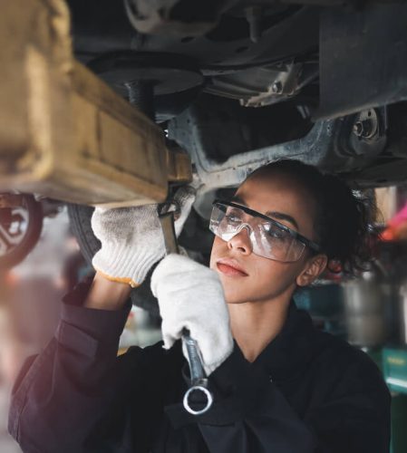 A woman working underneath a HGV holding a spanner and waring eye protection.