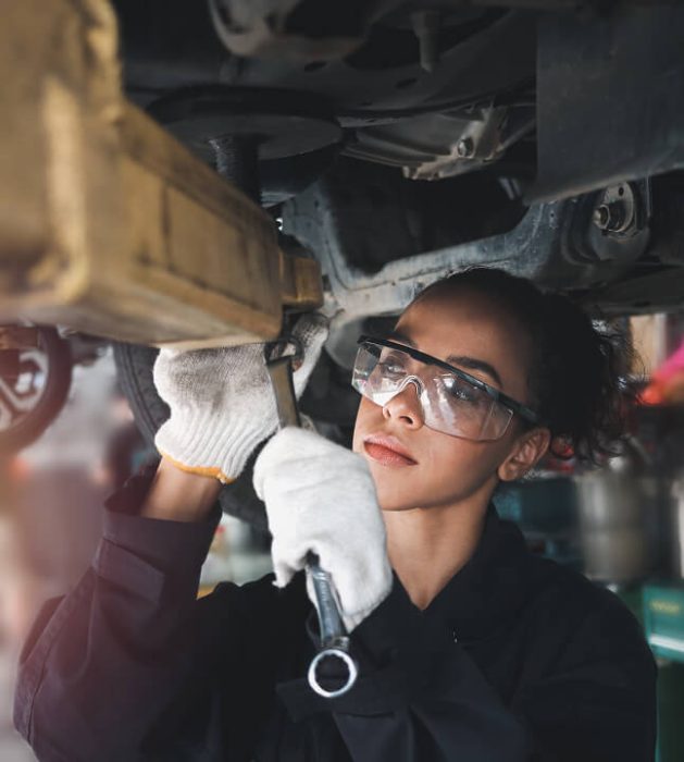 A woman working underneath a HGV holding a spanner and waring eye protection.