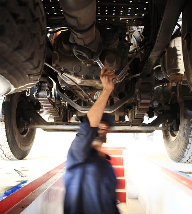 A HGV technician working underneath a large vehicle in blue overalls.