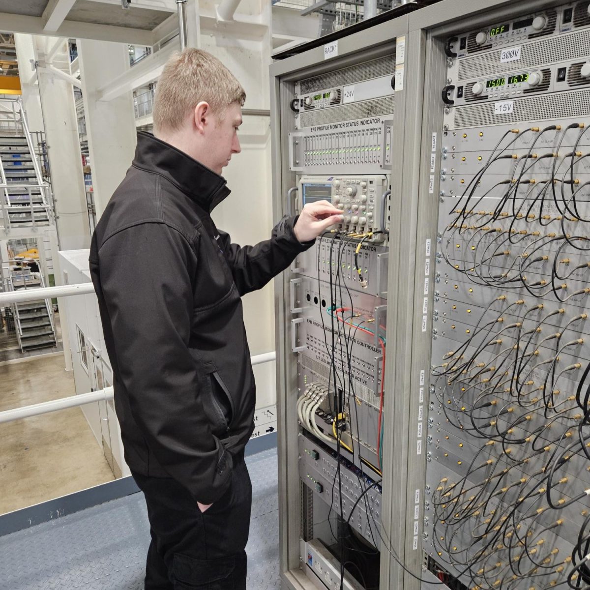 Electrical Engineering Technician Hayden, stood in front of a rack of electrical equipment, with wires spilling out.