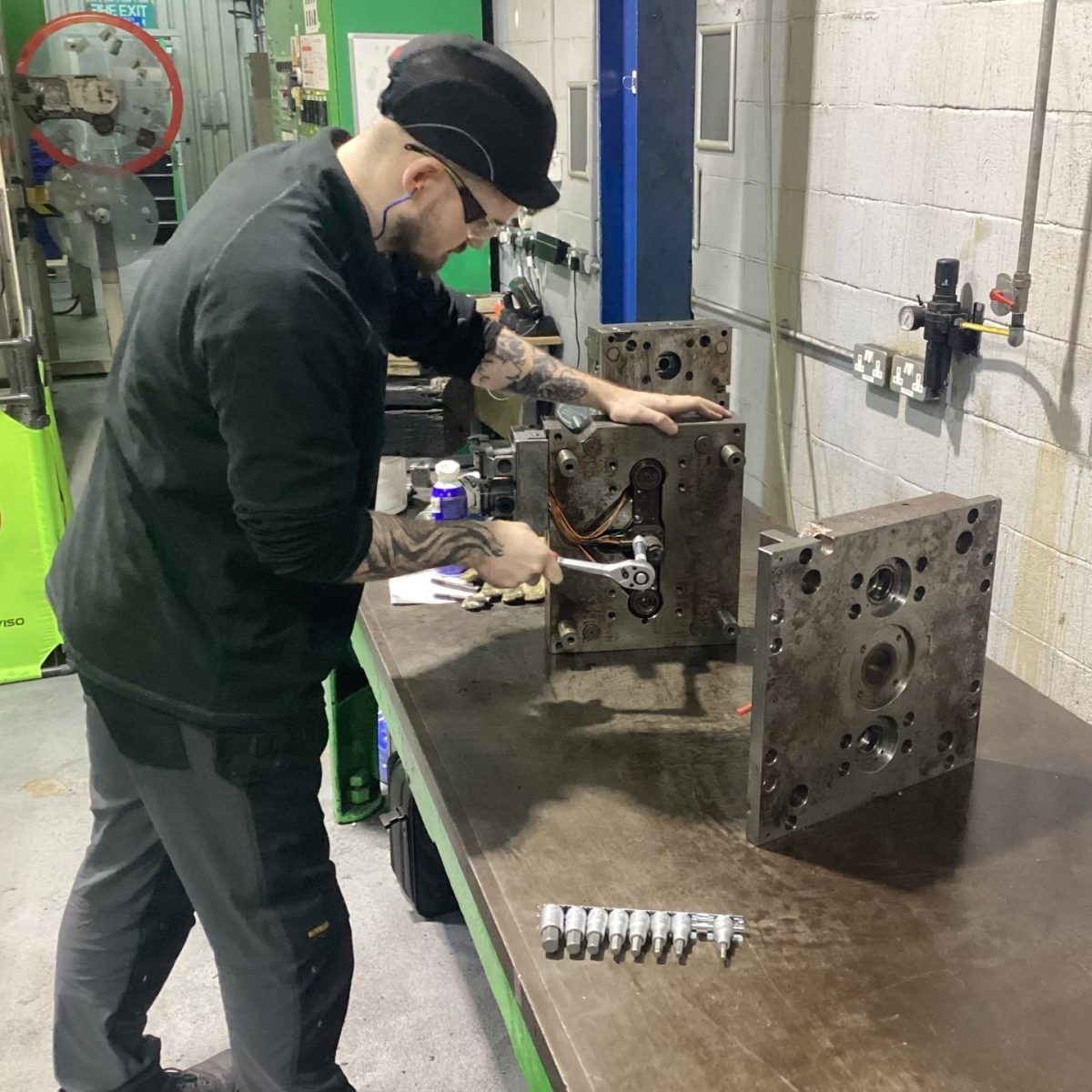 Maintenance and Operations Engineering Technician James, in the workshop, wearing safety goggles and hat, using a ratchet to fix a piece of equipment on the workbench.