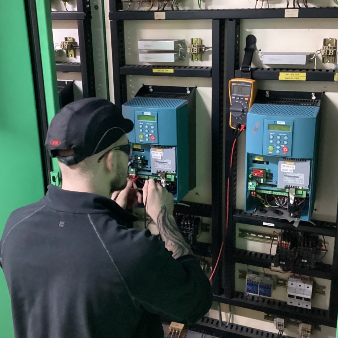Maintenance and Operations Engineering Technician James, wearing safety glasses and a hat, using a multimeter to test some electrical components on a wall.