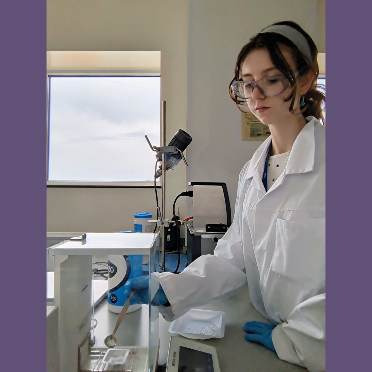 Research Laboratory Technician Jorja in the lab, wearing a lab coat and protective glasses, using a spatula to measure out and weigh raw materials.