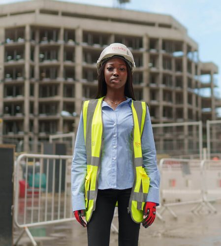 Woman in blue shit with a high-vis vest and hard hard stood in front of a construction site