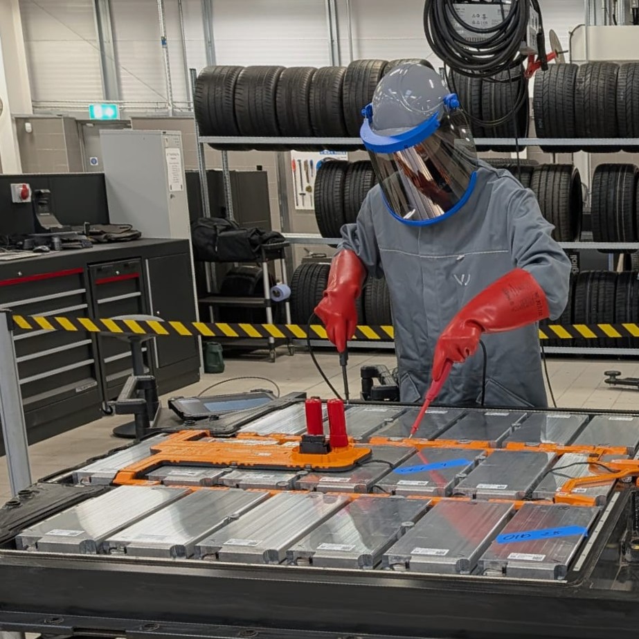 Electric Vehicle Technician Pete, in the garage, working with high voltage equipment on a tabletop, wearing a full arc flash suit, including face visor and insulated rubber gloves.