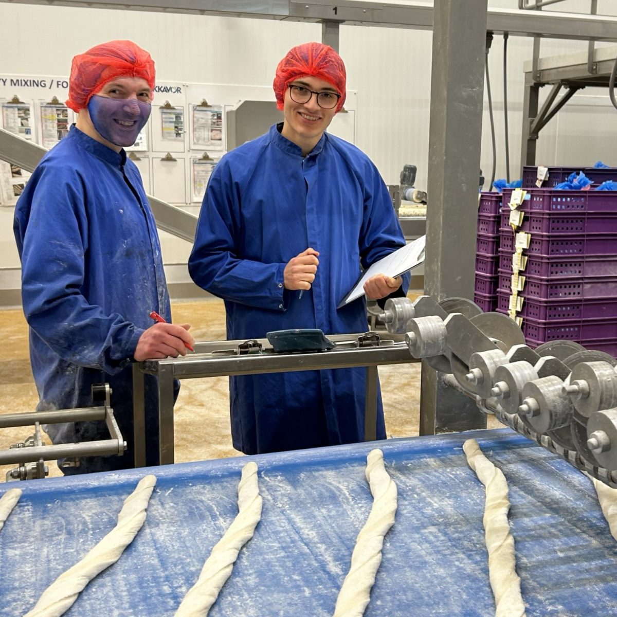 Food Science Technician William in the factory with a colleague, both wearing overalls and hair nets, standing next to rolls of fresh dough.