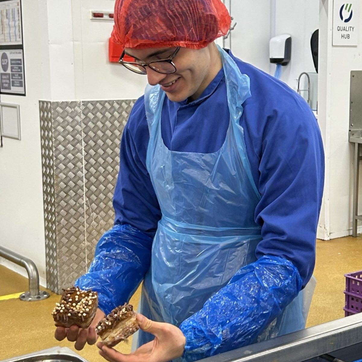 Food Science Technician William in the factory holding two pieces of cake, whilst wearing overalls, hair net, and a plastic apron.