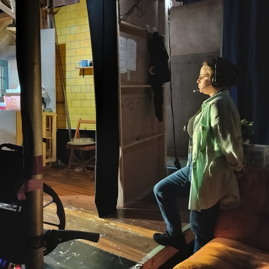 Creative Venue Technician Zoë, wearing a headset and mic, standing offstage, in the wings of a theatre show.