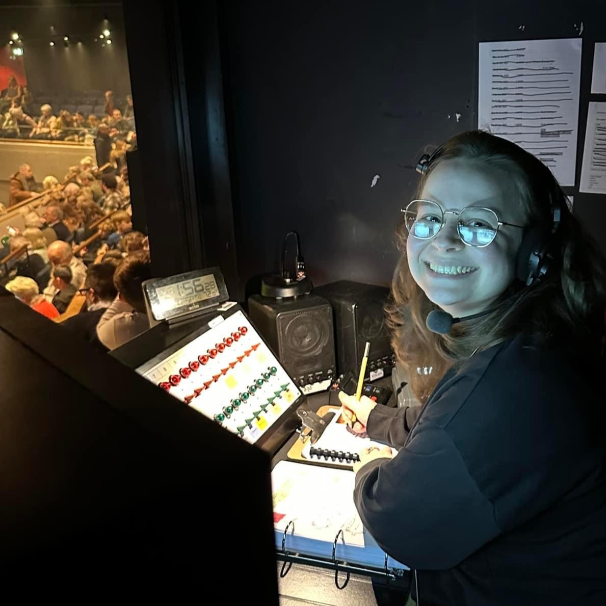 Creative Venue Technician Zoë, wearing a headset and mic, sat backstage at a control desk during a theatre show.