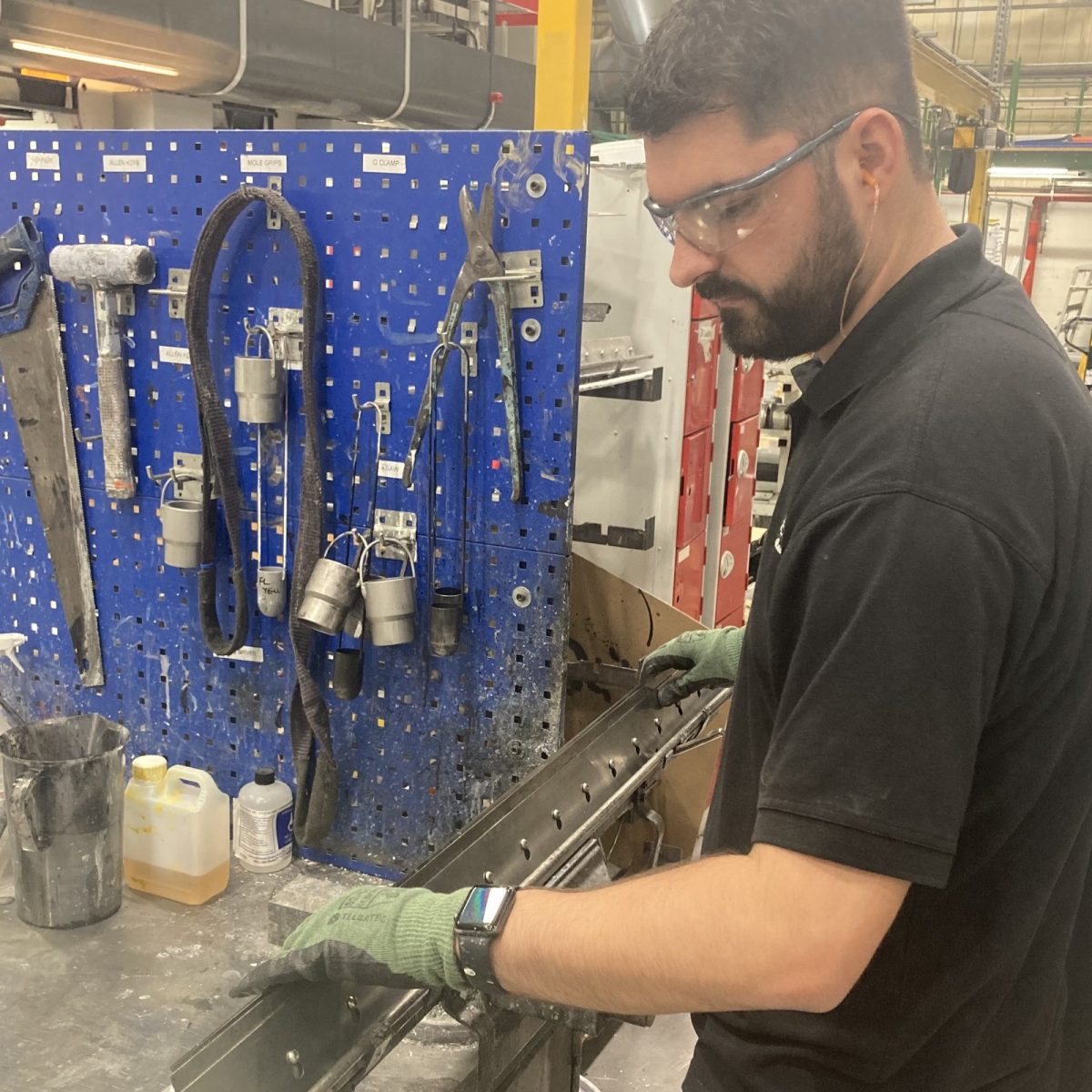 Print Technician Jack, wearing gloves, safety glasses, and ear plugs, working with a piece of metal on a workbench, with tools hanging off the wall.