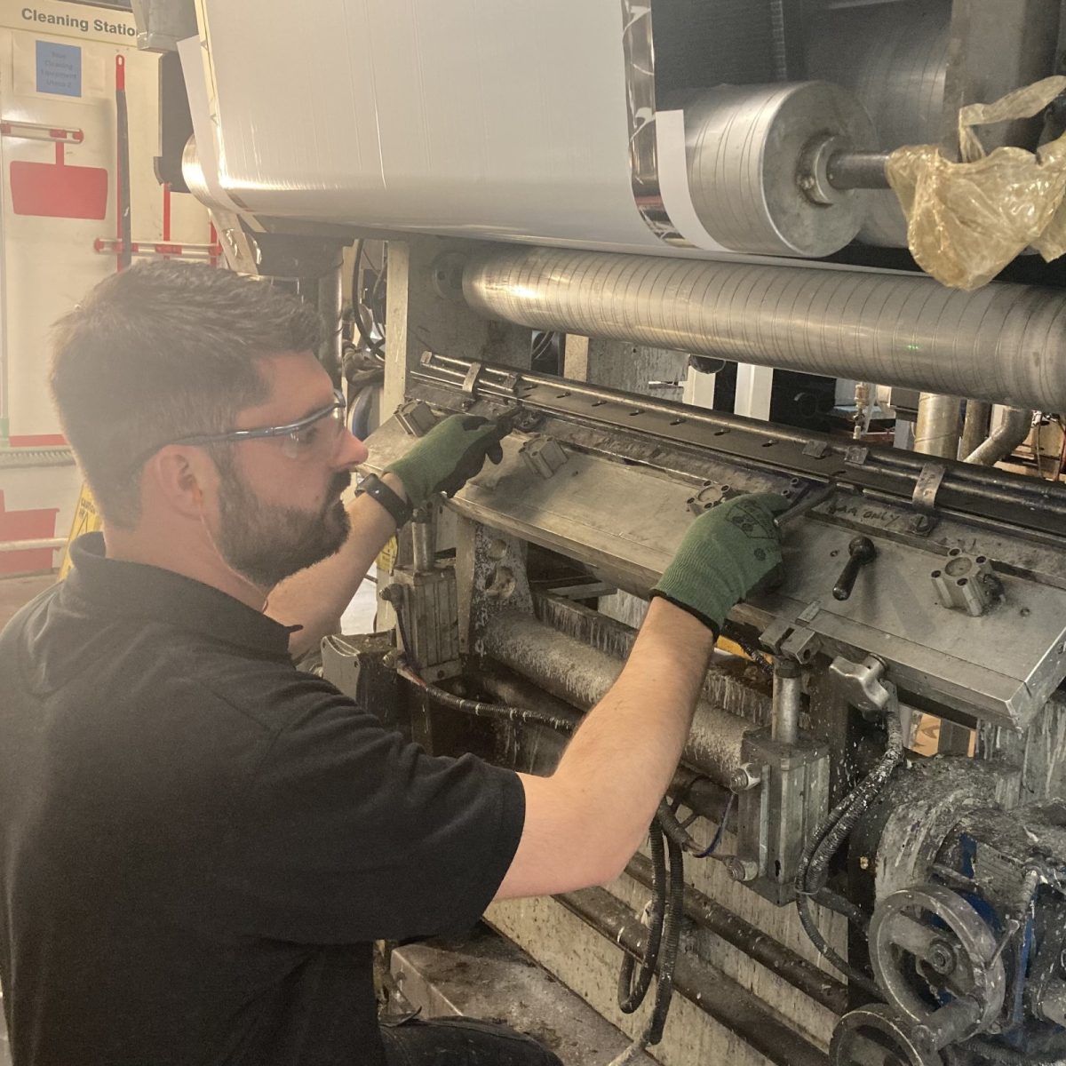 Print Technician Jack, wearing gloves, safety glasses, and ear plugs, working on a stopped and open printing machine.