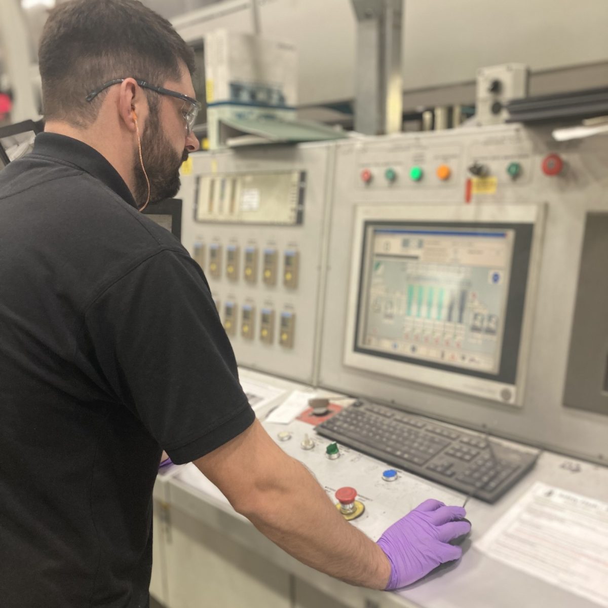 Print Technician Jack, wearing gloves, safety glasses, and ear plugs, working on a screen at a machine control desk.