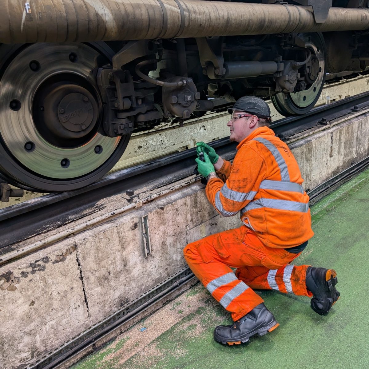 Rail Engineering Technician Josh, wearing hi-vis overalls, gloves, hard hat, and safety goggles, kneeling down next to a train's wheels on tracks in the depot.