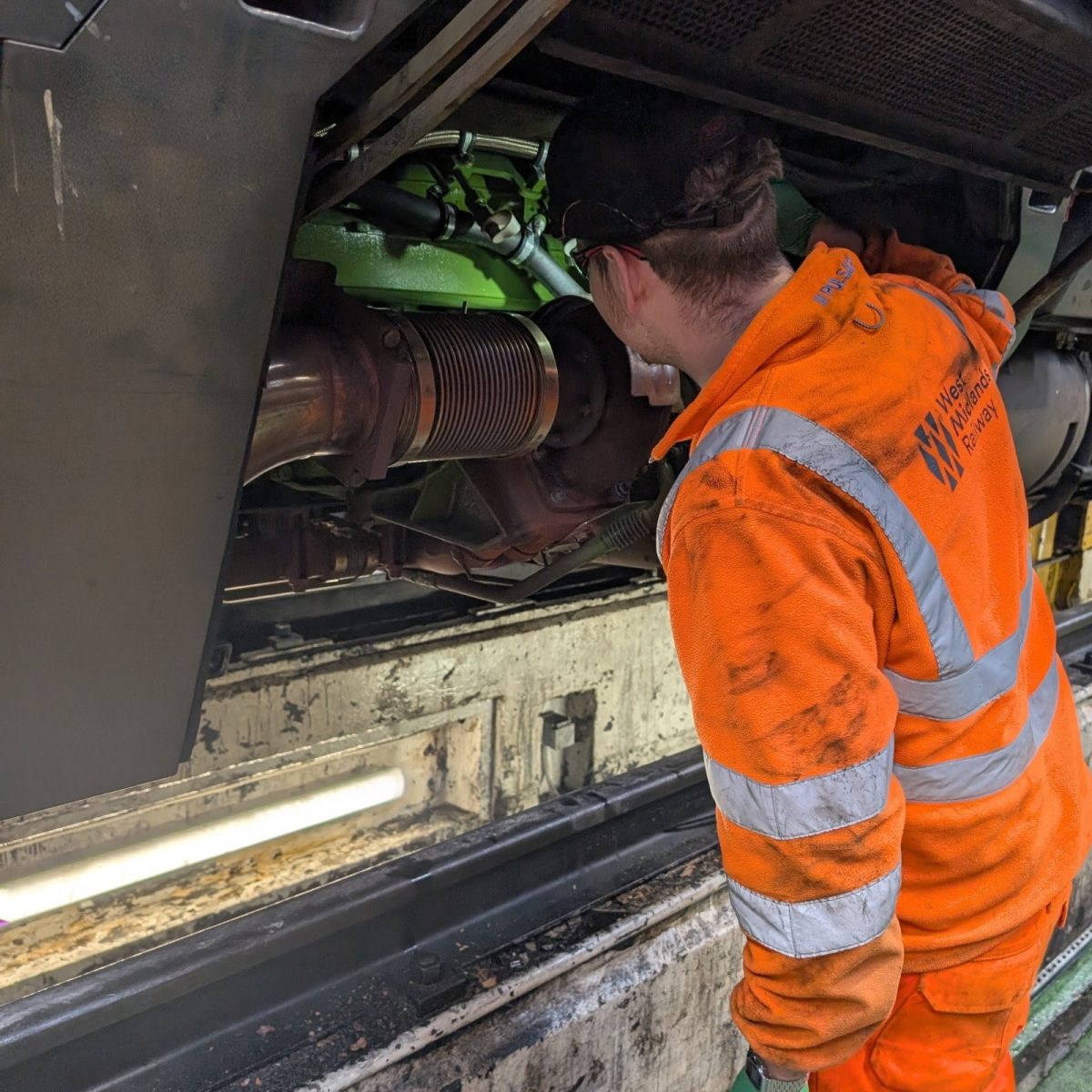 Rail Engineering Technician Josh, wearing hi-vis overalls, gloves, hard hat, and safety goggles, working on a train's undercarriage in the depot.