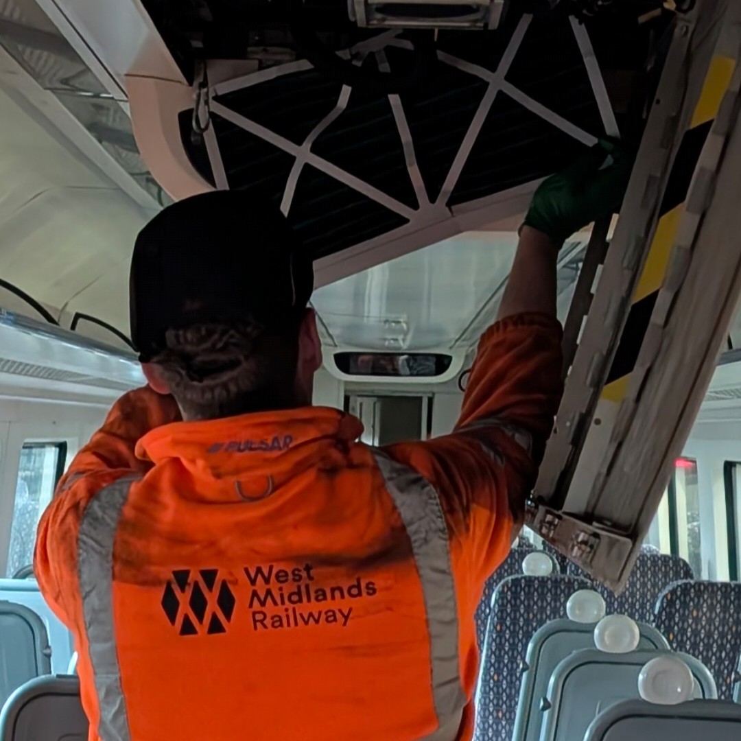 Rail Engineering Technician Josh, wearing hi-vis overalls, gloves, hard hat, and safety goggles, working on an open ceiling compartment, in a passenger carriage whilst the train is in the depot.