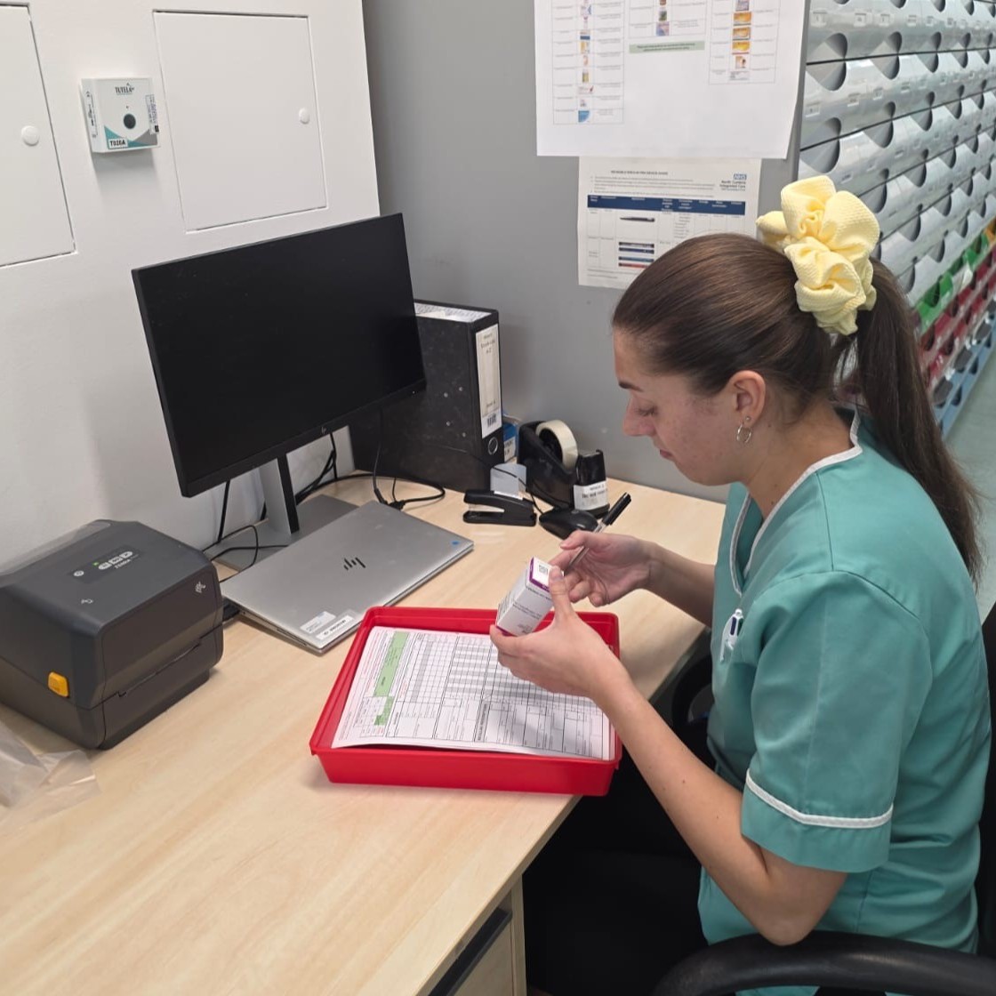 Pharmacy Technician Megan, wearing a healthcare tunic, sat at a desk, with a packed of medicine, filling in some paperwork.
