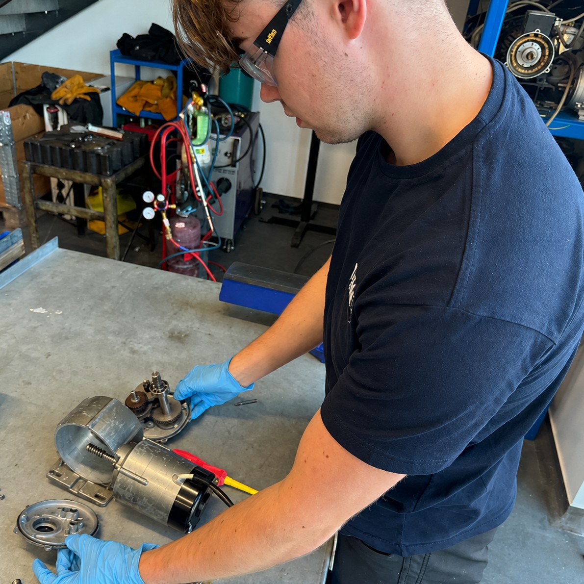 Wind Turbine Technician Munro, wearing safety glasses and blue protective gloves, working at a metal workbench, handling an exposed electric motor and mechanical components. There are tools, cables, and industrial equipment in the workshop background.