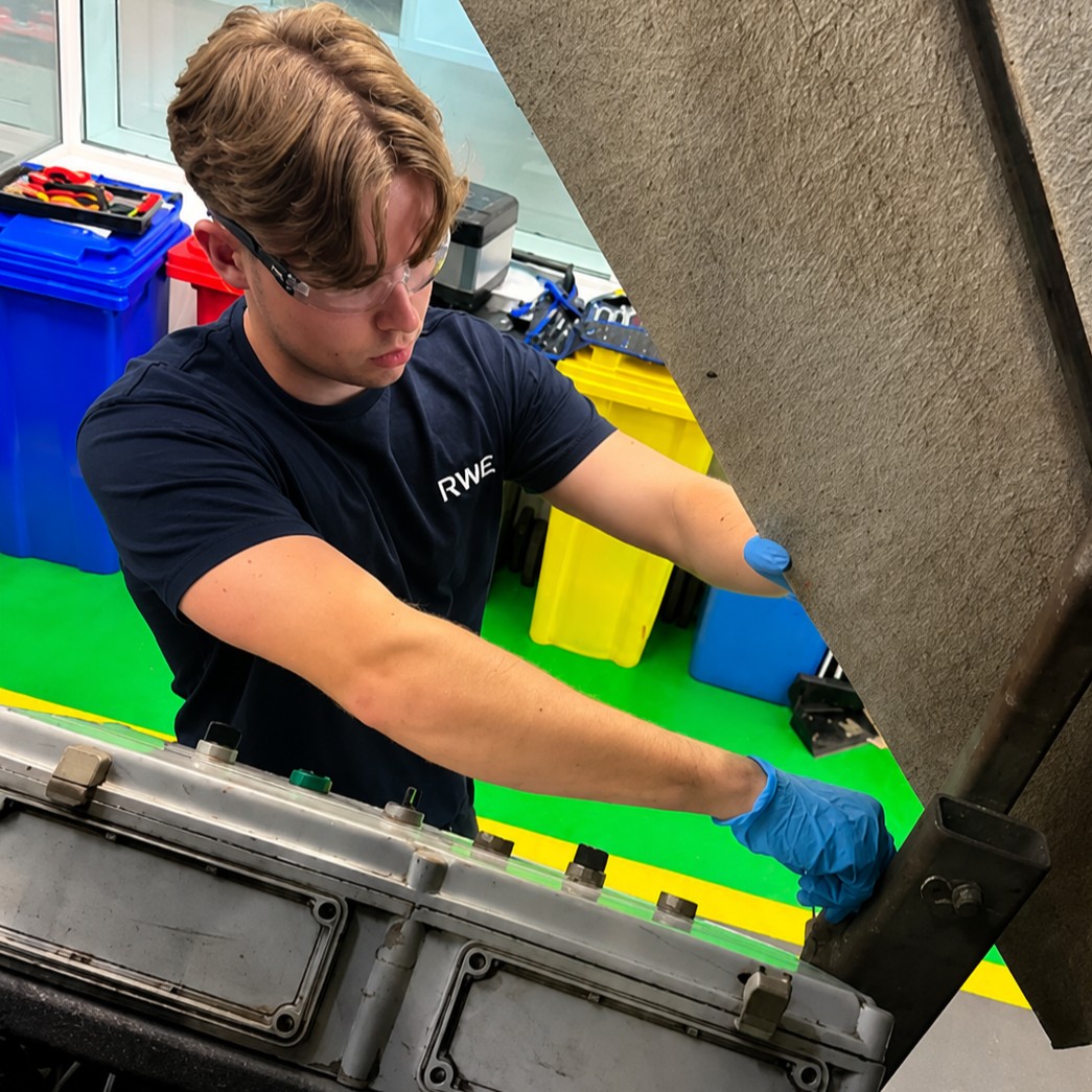 Wind Turbine Technician Munro, wearing latex gloves and safety glasses, working inside industrial equipment – reaching into an open machine housing in a workshop with colour-coded recycling bins in the background.
