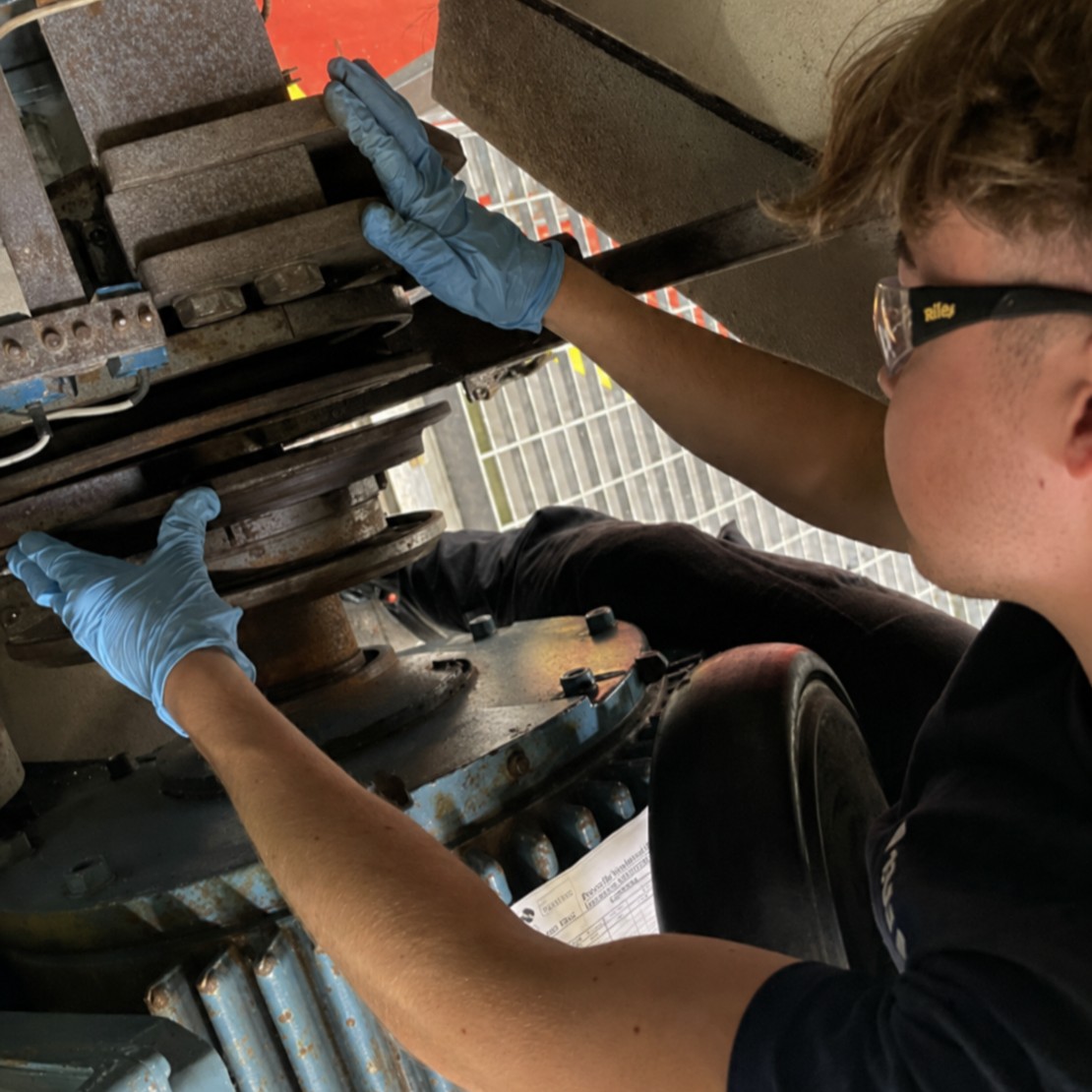Wind Turbine Technician Munro, wearing safety glasses and blue gloves, inspecting heavy mechanical parts inside a large industrial machine.