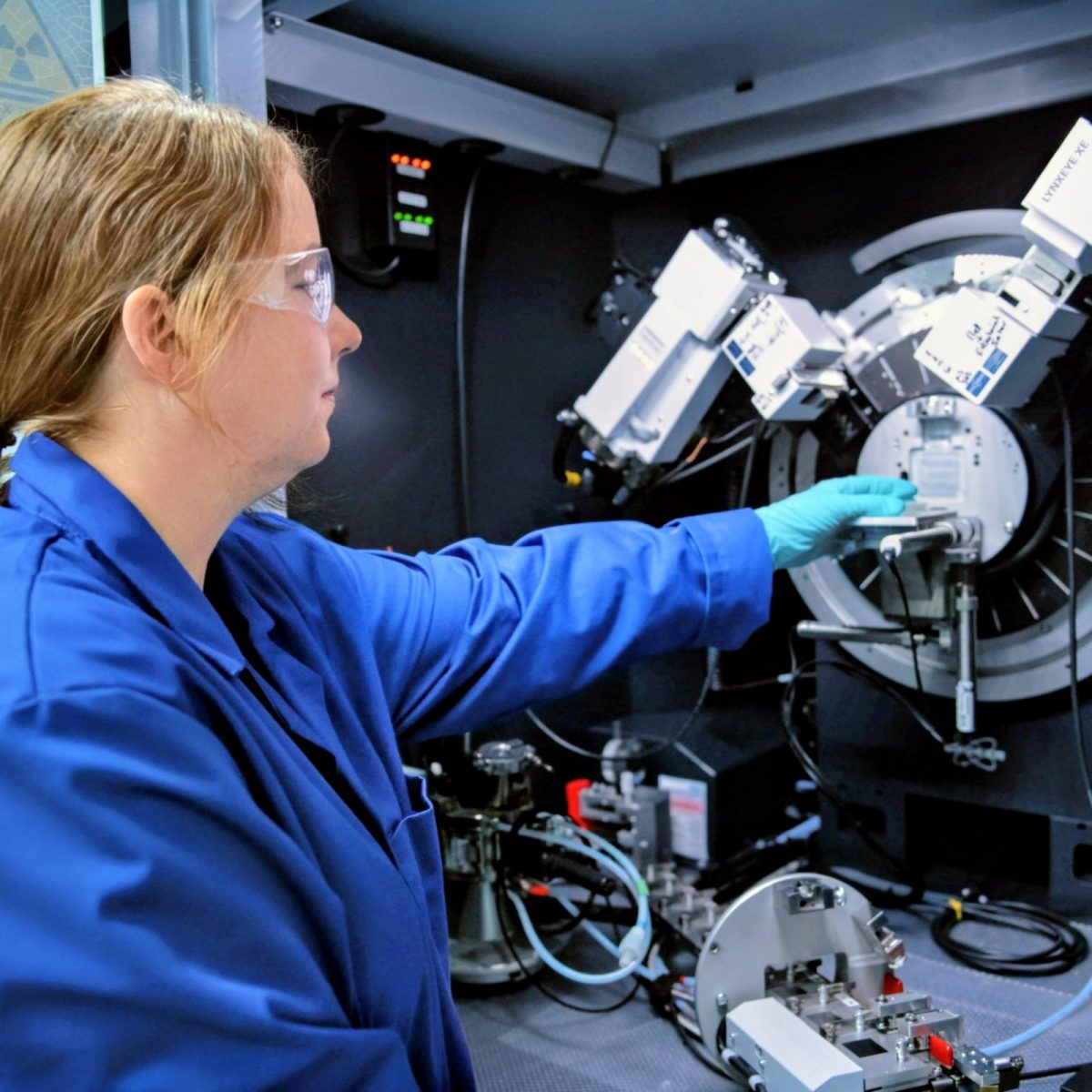 Testing Engineering Technician Sophie, wearing a lab coat, glove, and safety glasses, positioning a sample inside analytical equipment in a laboratory.