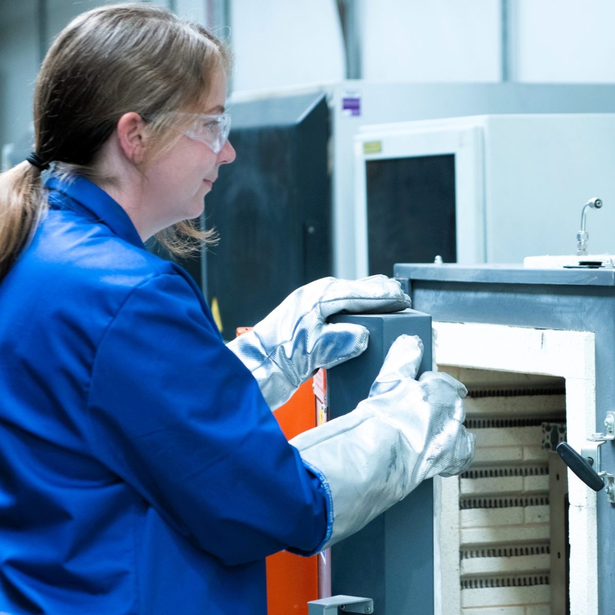 Testing Engineering Technician Sophie, wearing a lab coat, heat-resistant gloves, and safety glasses, opening a laboratory furnace.
