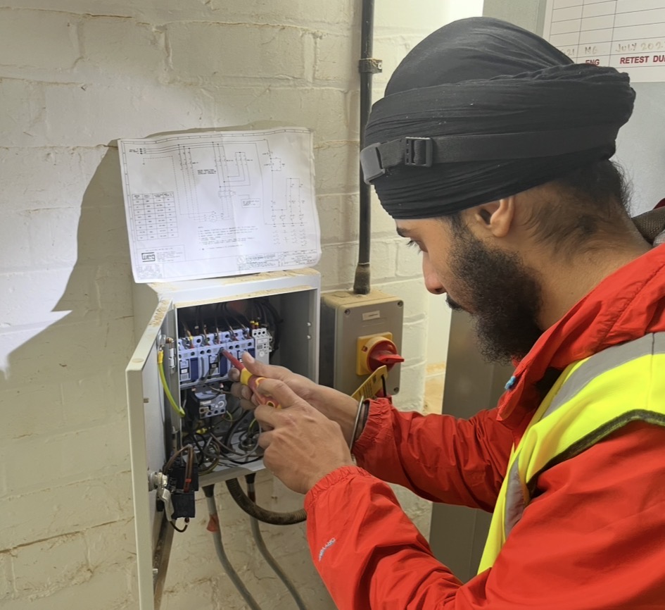 Electrical Engineering Technician Tanvir, wearing a hi-vis jacket and headtorch, adjusting an open electrical junction box.