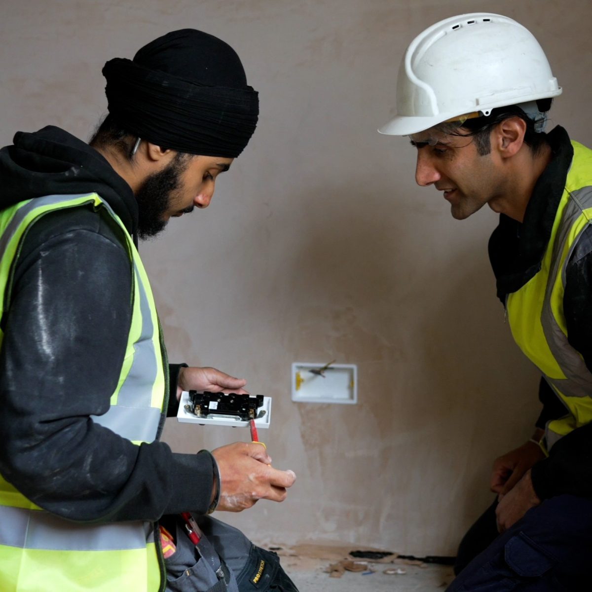 Electrical Engineering Technician Tanvir, wearing a hi-vis jacket, inspecting an electrical component with a colleague wearing a hardhat.