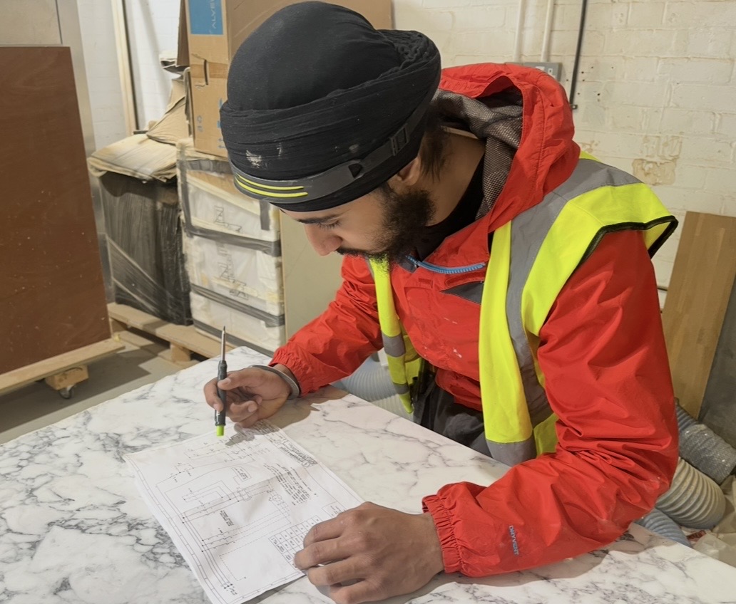 Electrical Engineering Technician Tanvir, wearing a hi-vis jacket, inspecting a wiring diagram on a countertop.