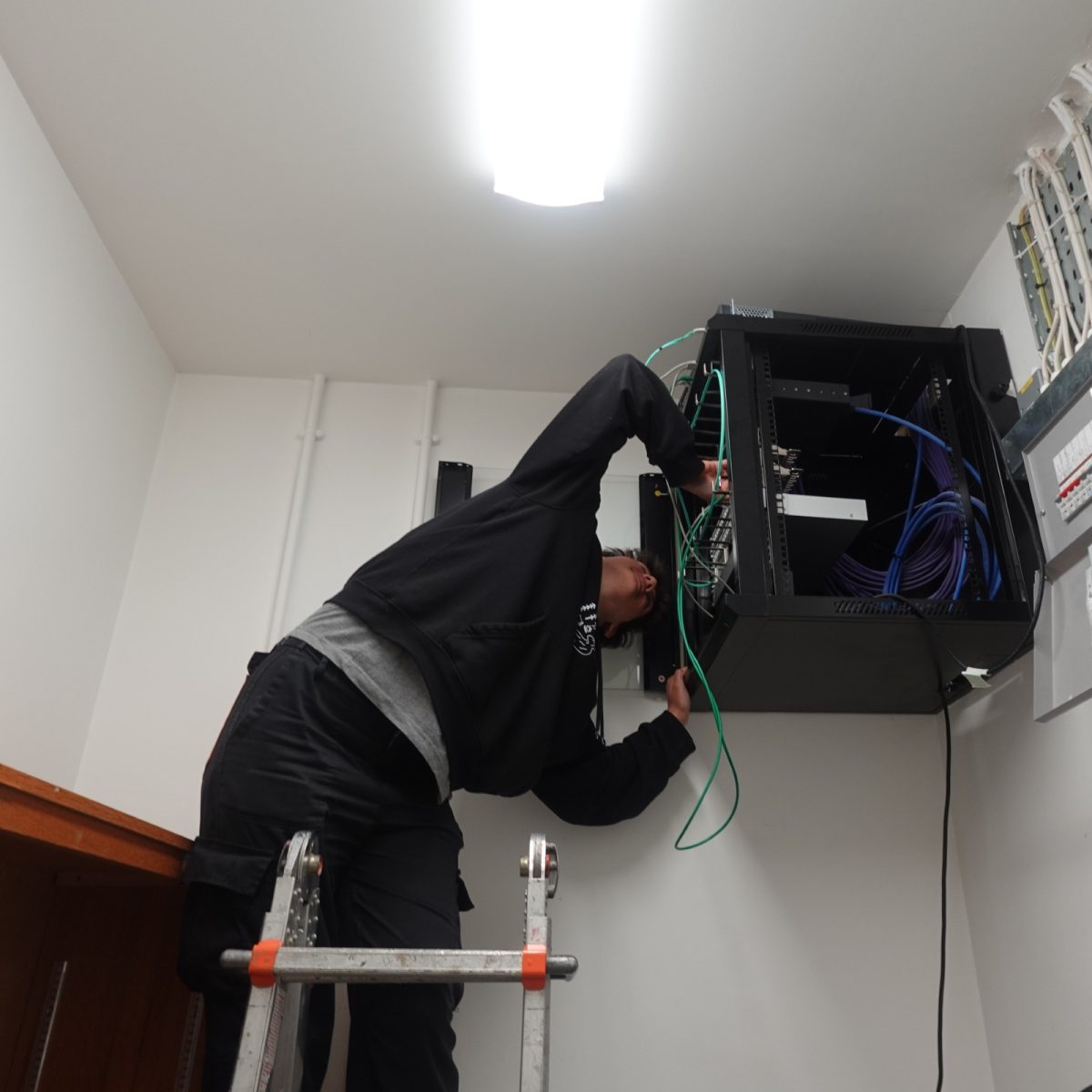 Software Developer Jaz, wearing a hoodie and standing on a ladder, working with network cables in a wall-mounted server cabinet.