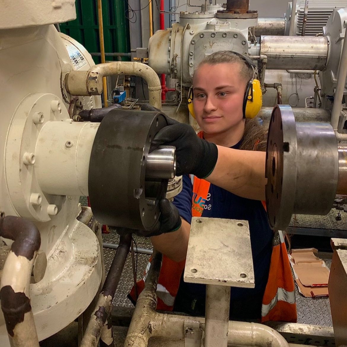 Refrigeration Engineering Technician Millie, wearing protective gloves, ear defenders, and a hi-vis vest, fixing large industrial machinery.