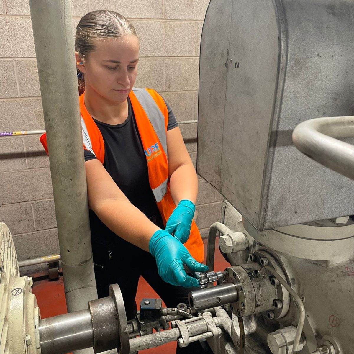 Refrigeration Engineering Technician Millie, wearing protective gloves, ear defenders, and a hi-vis vest, fixing large industrial machinery with a hand tool.