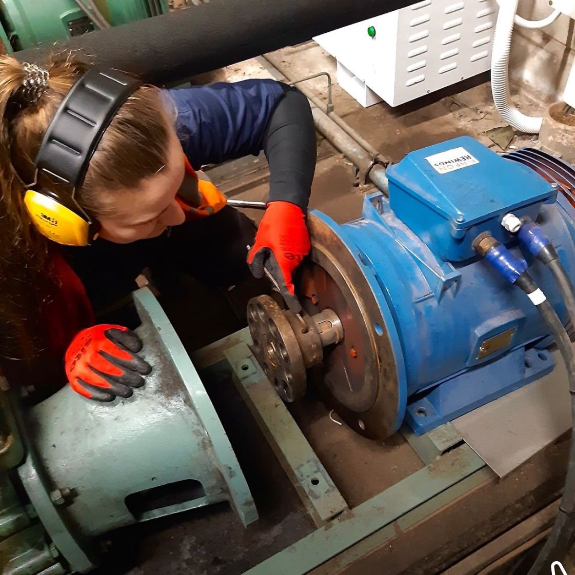 Refrigeration Engineering Technician Millie, wearing protective gloves, ear defenders, and a hi-vis vest, fixing large industrial machinery with a hand tool.