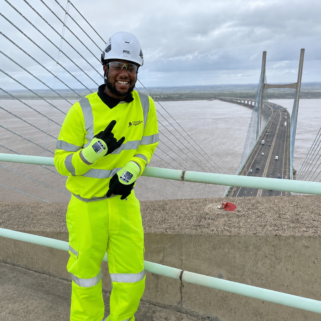 Civil Engineering Technician Tochi, wearing a full hi-vis outfit, protective gloves, safety goggles, and a hard hat, smiling and posing on top of the River Severn Suspension Bridge, with cars driving over the bridge below.