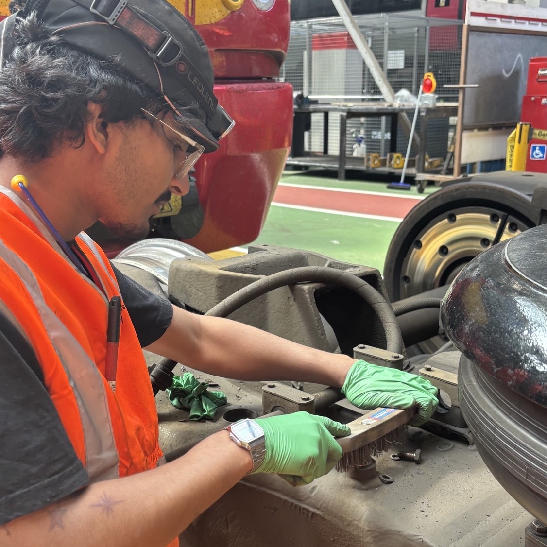 Railway Engineering Technician Dev, wearing a hi-vis vest, gloves, safety glasses, and a cap, cleaning a train component with a wire brush, in a rail depot workshop.